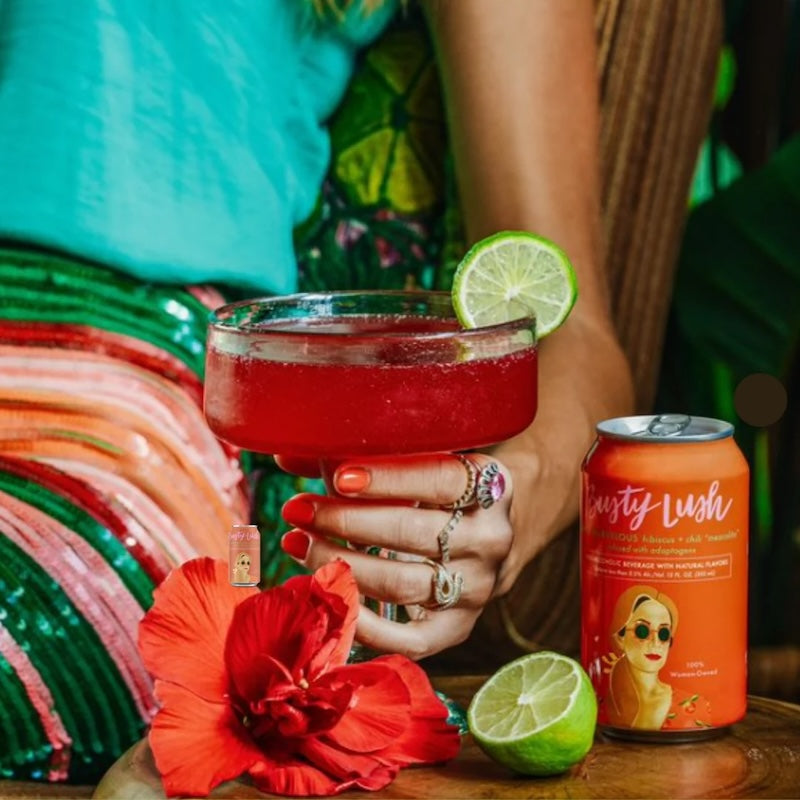 A person holding a glass of red mezcalita with a slice of lime on the rim, next to a can of Busty Lush She's Marvelous Hibiscus + Chili Mezcalita. There is also an orange with a hibiscus flower and a colorful patterned fabric in the background.
