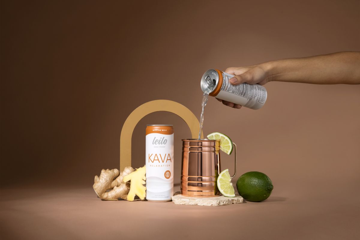 Person pouring a drink into a copper mug with Kava can and ingredients on a brown background