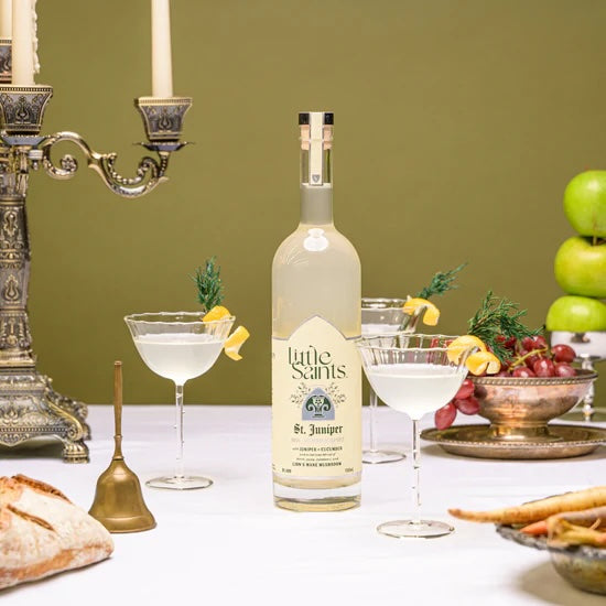 A bottle of Little Saints Saint Juniper spirit, with two filled cocktail glasses and a partial glass, arranged on a table with a green background. Decorative items, such as a candle holder, and fruits are also visible in the setting.