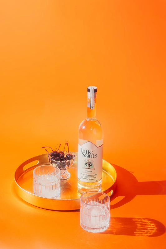 A bottle of gin with the label 'Saint Ember' is displayed on a golden tray, accompanied by two filled glasses and a bowl of olives, against an orange background.