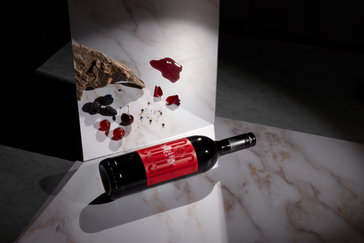 A bottle of Noughty Alcohol-Free Rouge Syrah wine lying on a marble surface with a decorative rose petal arrangement and a piece of charcoal artfully placed in the background.