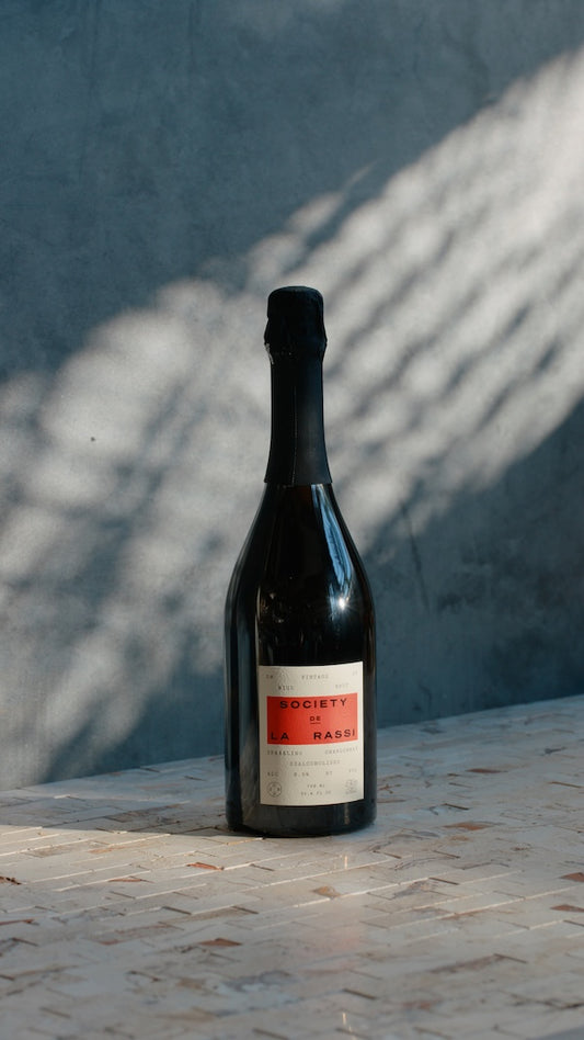 A bottle of Society de la Rassi Neue Brut champagne with a shadow cast on its label, placed on a tiled surface.