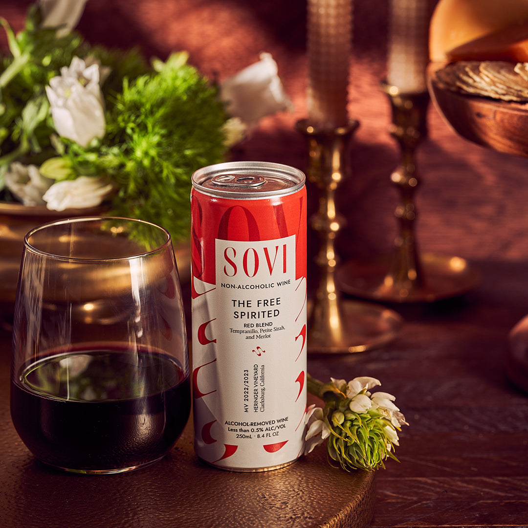 A can of Sovi non-alcoholic red blend wine with a glass of red wine and decorative candles in the background.