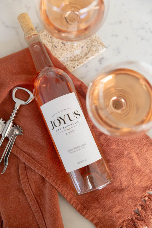 A bottle of Joyus Non-Alcoholic Rosé wine with two filled glasses beside it, set on a table with a cork screw and a napkin.