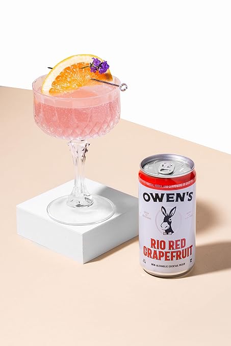 Cocktail in a glass with a slice of orange and purple flower, next to a can of Owen's Rio Red Grapefruit soda on a beige background.