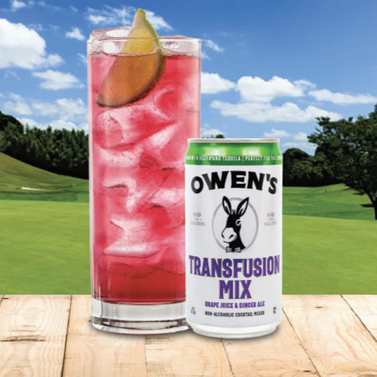 Glass of pink cocktail with a lime wedge and can of Owen's Transfusion Mix on a wooden surface with a golf course background.