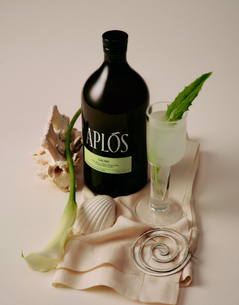 A bottle of Aplós Calme spiritless beverage with infused botanicals, displayed alongside a filled glass garnished with a green herb, a piece of citrus, and a shell on a cloth napkin.
