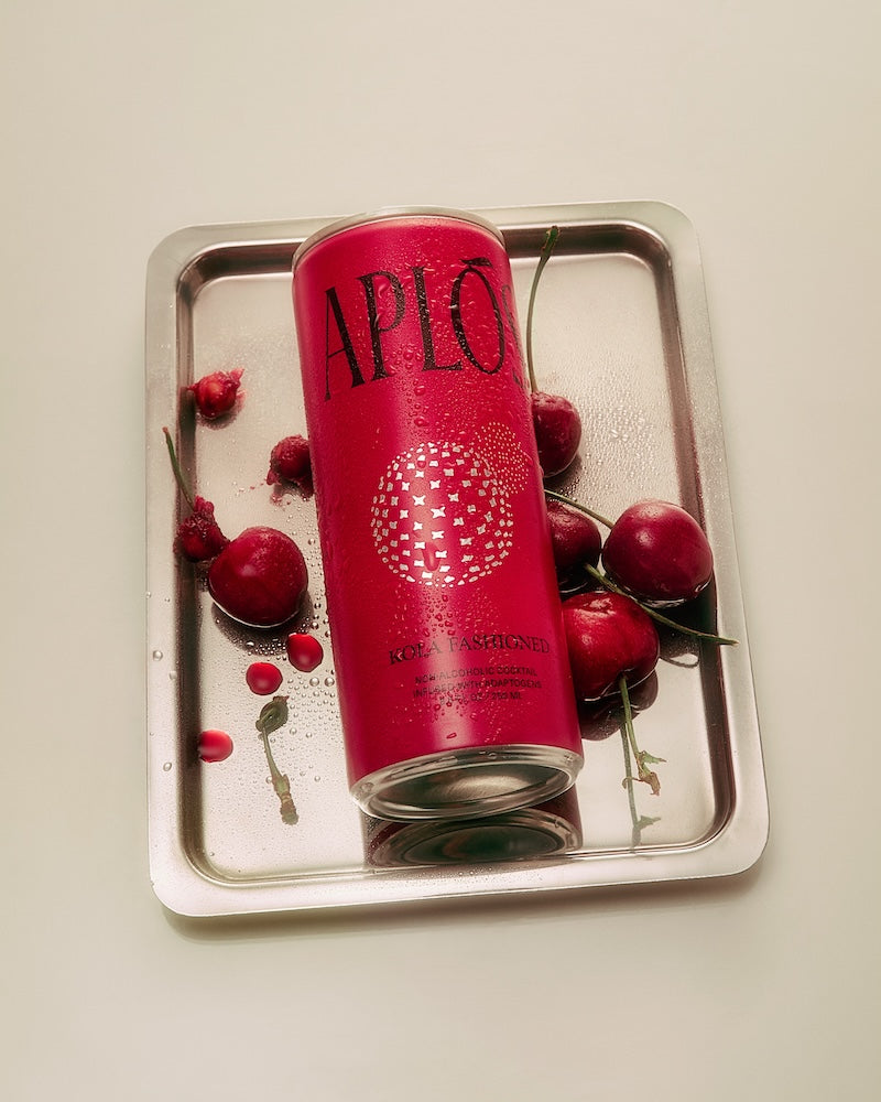 A can of Aplós Kola Fashioned cocktail placed on a tray with cherries around it, with the branding 'APLO' visible on the can.