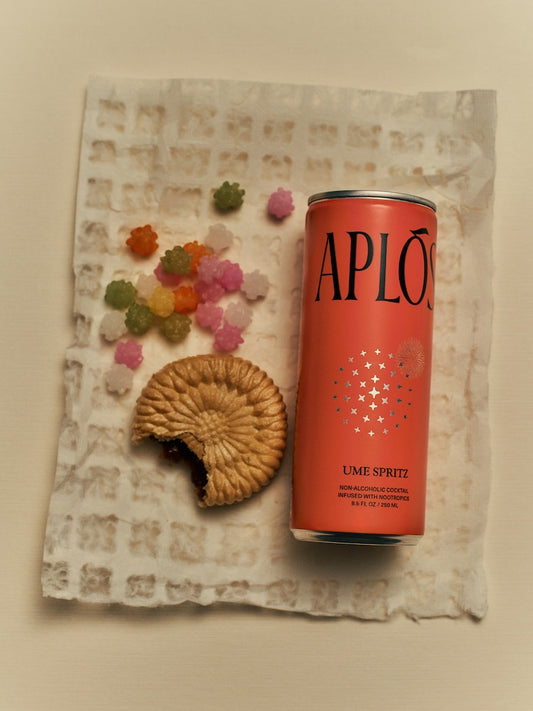 An image featuring a can of 'APLOS' Mandora Negroni sparkling, non-alcoholic cocktail, surrounded by colorful, decorative flowers and ice cubes. A cookie is also visible to the side.