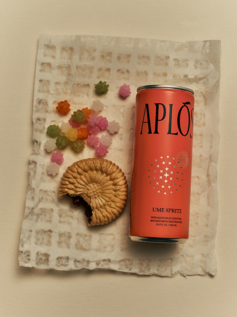 An image featuring a can of 'APLOS' Mandora Negroni sparkling, non-alcoholic cocktail, surrounded by colorful, decorative flowers and ice cubes. A cookie is also visible to the side.
