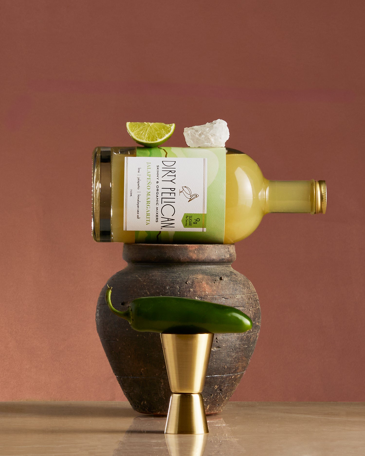 Bottle of 'Dirty Feliz' tequila with a lime wedge and jalapeΓ±o on a wooden and stone surface.