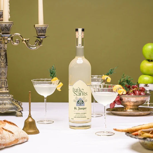 A bottle of Little Saints Saint Juniper spirit, with two filled cocktail glasses and a partial glass, arranged on a table with a green background. Decorative items, such as a candle holder, and fruits are also visible in the setting.