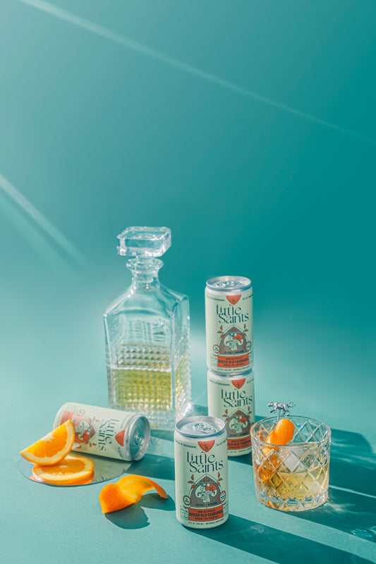 A 4-pack of Little Saints Spiced Old Fashioned cocktail mixes, with packaging that resembles a vintage apothecary. One can is open, revealing the cocktail inside, and a second can is visible with an orange slice and a lemon slice beside it, implying the flavor of the cocktail.