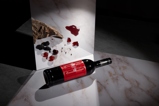 A bottle of Noughty Alcohol-Free Rouge Syrah wine lying on a marble surface with a decorative rose petal arrangement and a piece of charcoal artfully placed in the background.