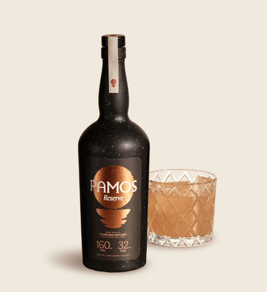 A bottle of Pamos Reserve spirit next to a glass with a geometric pattern. The bottle is dark colored with a label that includes the brand name and 'Reserve' written on it.