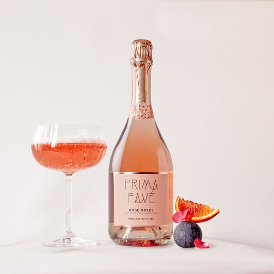 A bottle of Prima Pave Rose Dolce with a glass filled with pink sparkling wine, alongside sliced fruits and a flower.