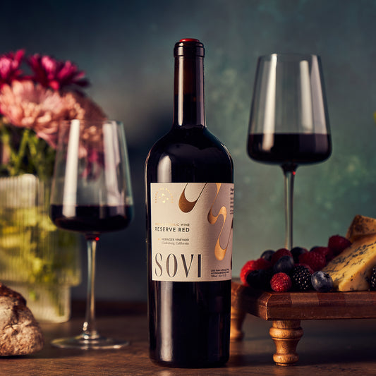 A bottle of Sovi Reserve Red wine is displayed in front of two filled wine glasses. In the background, there are flowers and a variety of cheeses arranged on a wooden board.