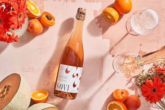 The image features a bottle of Sovi The Wild Child white wine, positioned prominently amidst a vibrant assortment of fresh citrus fruits including oranges, mandarins, and pears, as well as an array of colorful flowers and a glass of bubbling champagne, suggesting a sophisticated and fruity theme for the product.