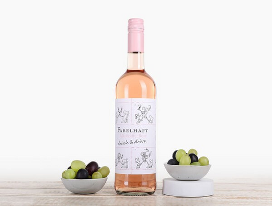 A bottle of Fabelhaft Alcohol-free Rosé wine with a pink label, accompanied by bowls of dark and green grapes on a wooden surface.